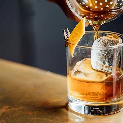 A whiskey or similar cocktail on the rocks, garnished with an orange twist and a large ice cube, poured into a clear tumbler on a bar.