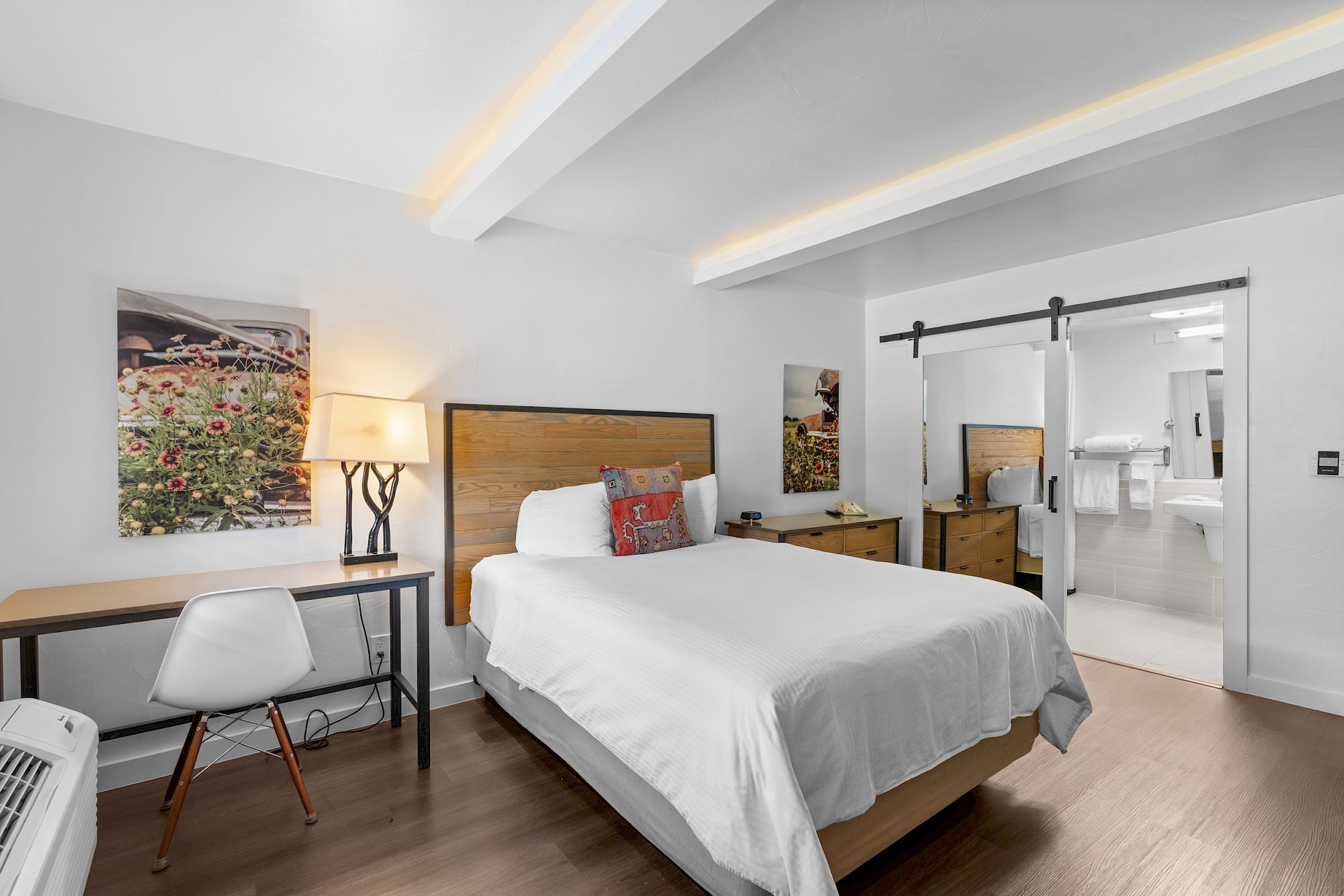 A modern hotel room with a queen bed, white linens, a wooden headboard, a desk and chair, a floor lamp, wall art, and a sliding barn door leading to a bathroom, all in light, airy decor.