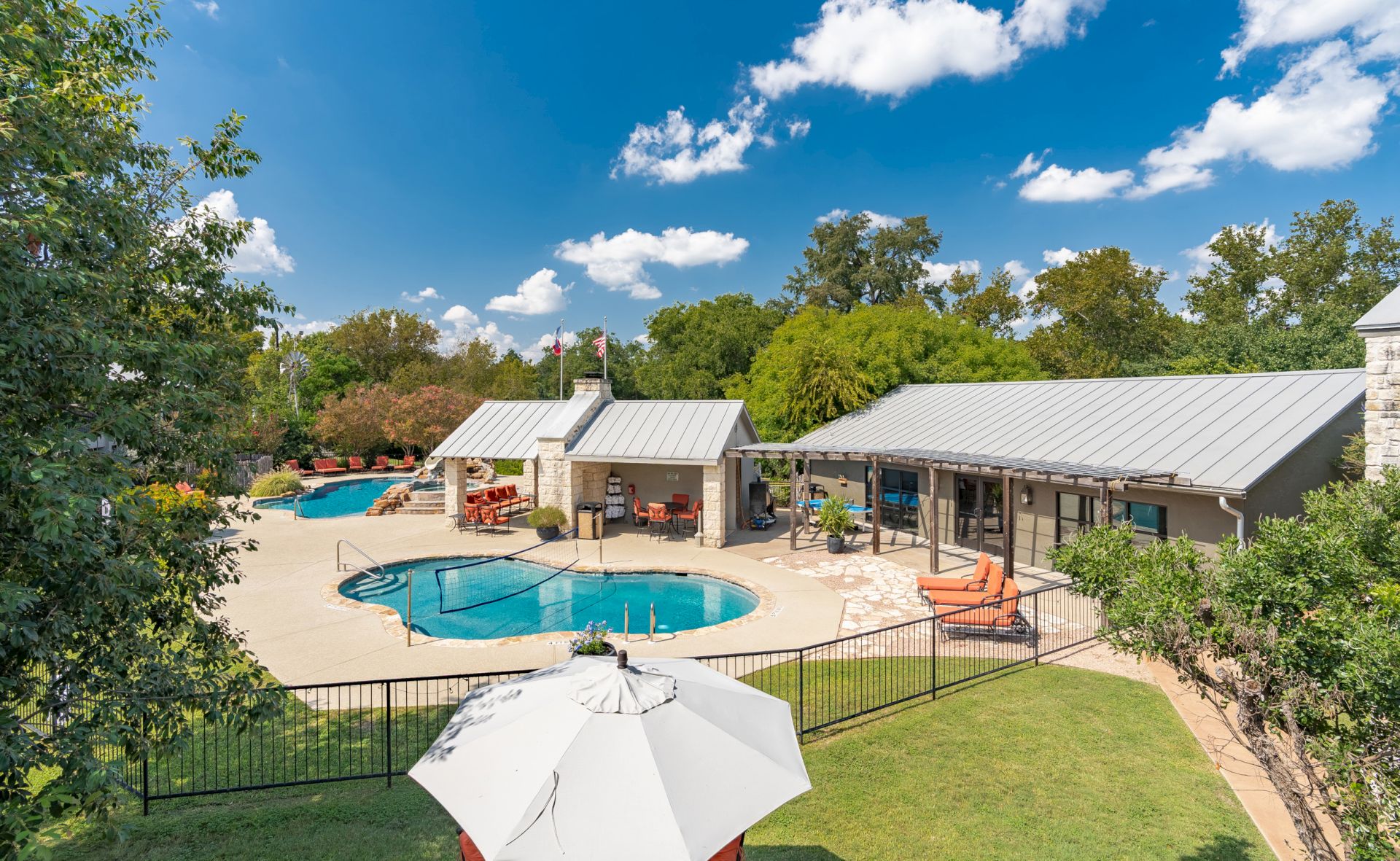The image shows a backyard with a swimming pool, buildings with metal roofs, patio furniture, and a large umbrella under a blue sky.