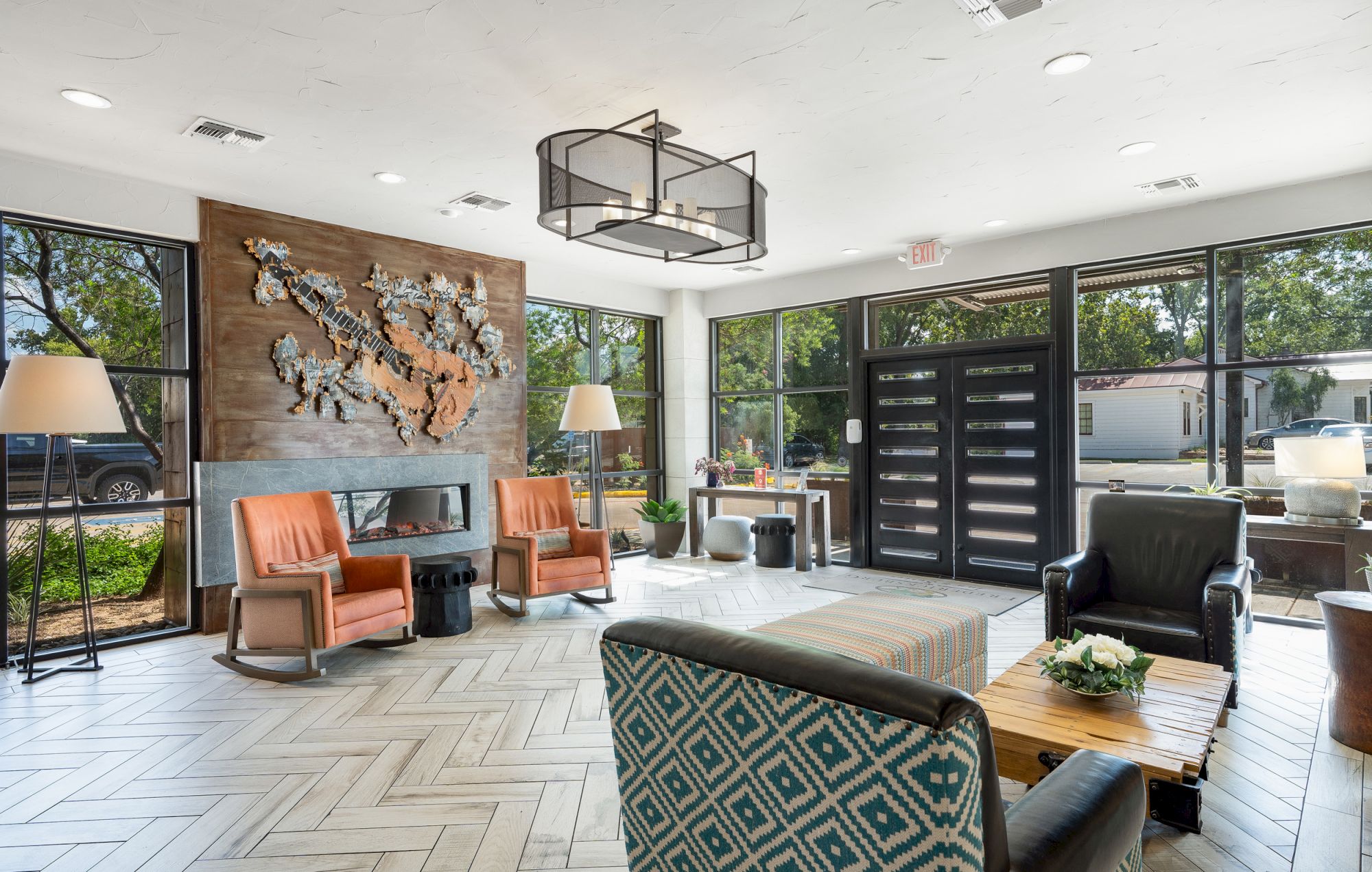 A modern lobby with armchairs, a decorative wall piece, large windows, and a central table with a plant, offering a welcoming ambiance.
