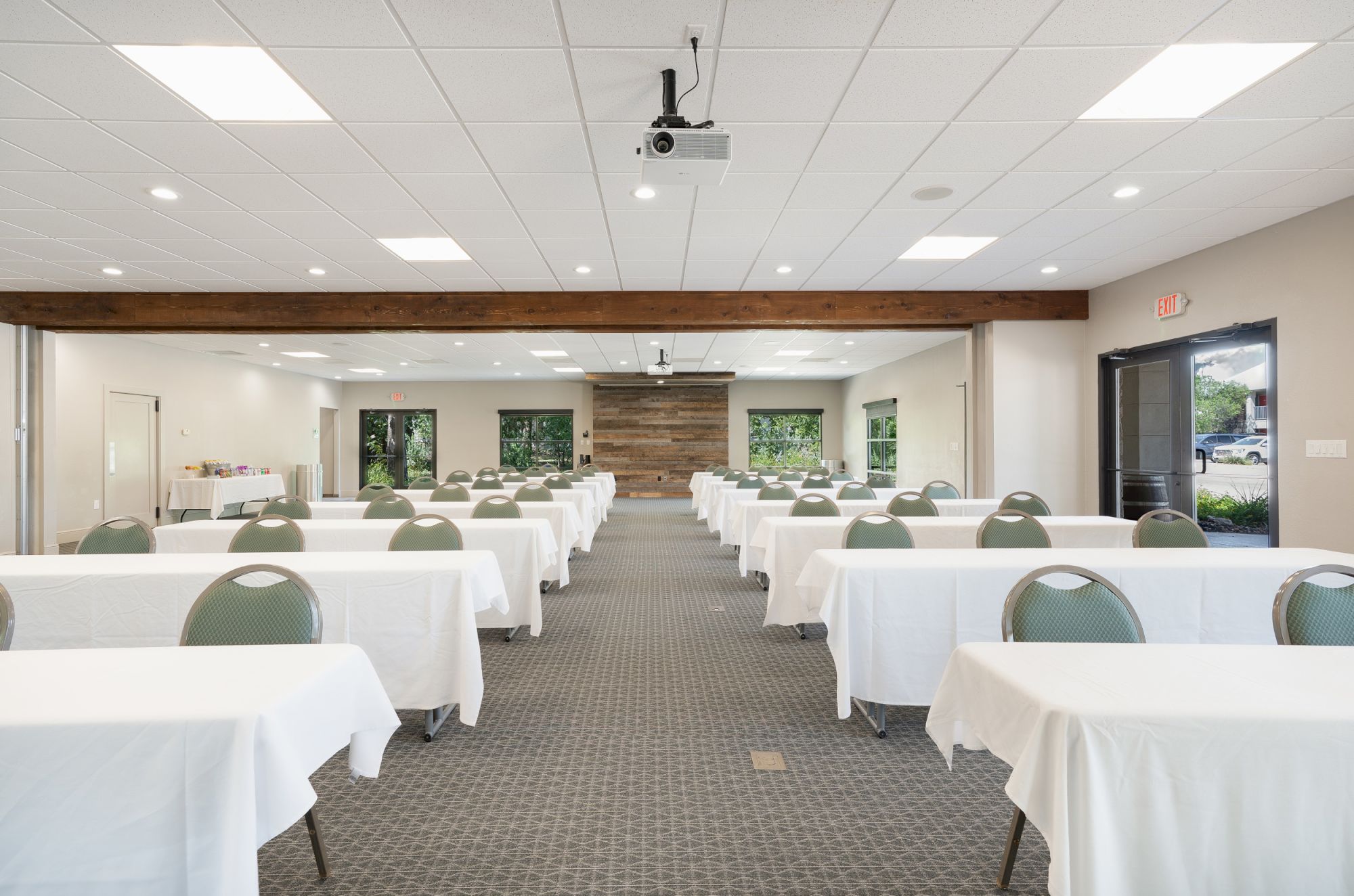 A conference room with rows of tables and chairs, each covered with white tablecloths, and a projector mounted on the ceiling.