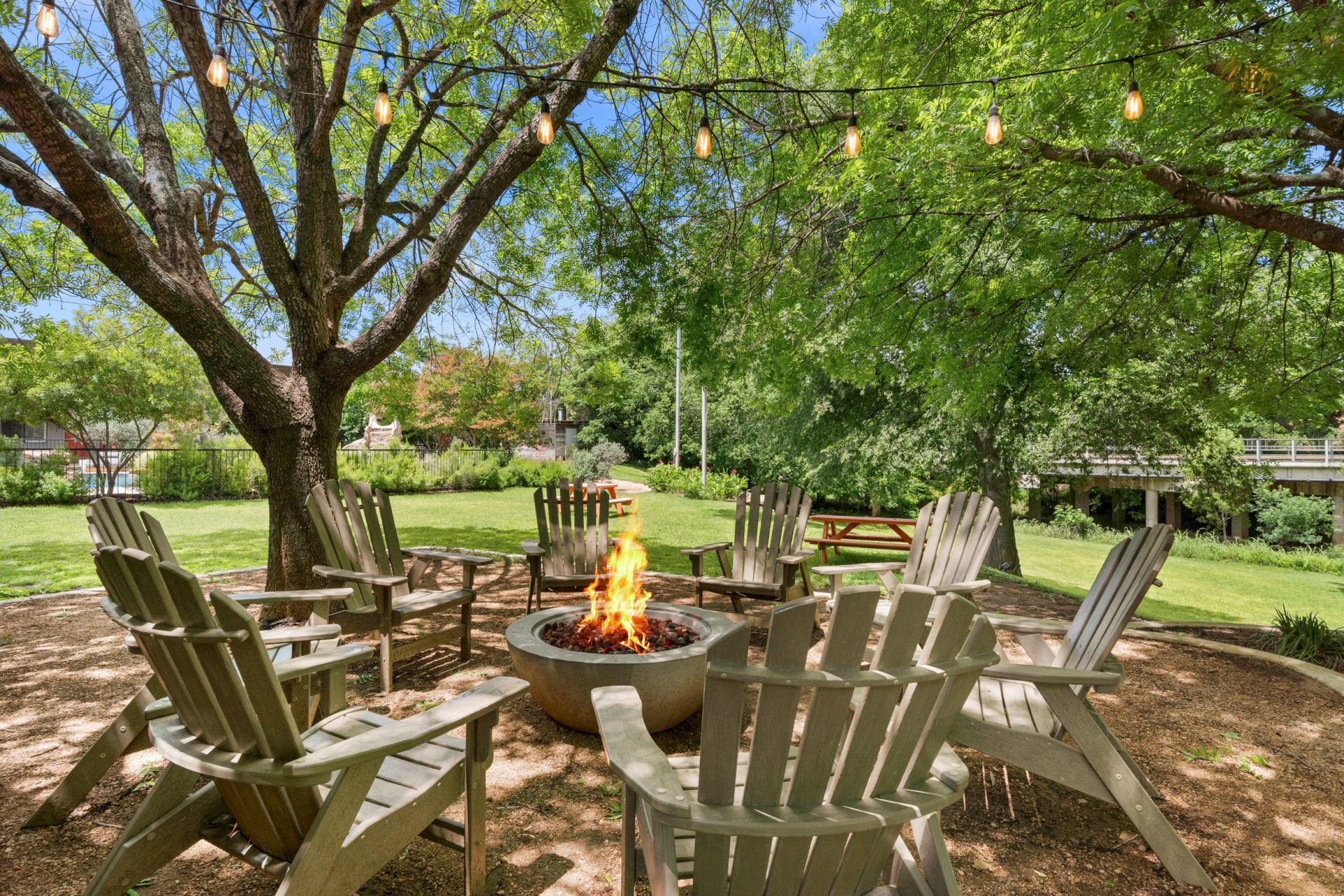 A cozy outdoor fire pit surrounded by wooden chairs under trees with string lights, perfect for a relaxing gathering by the flames.