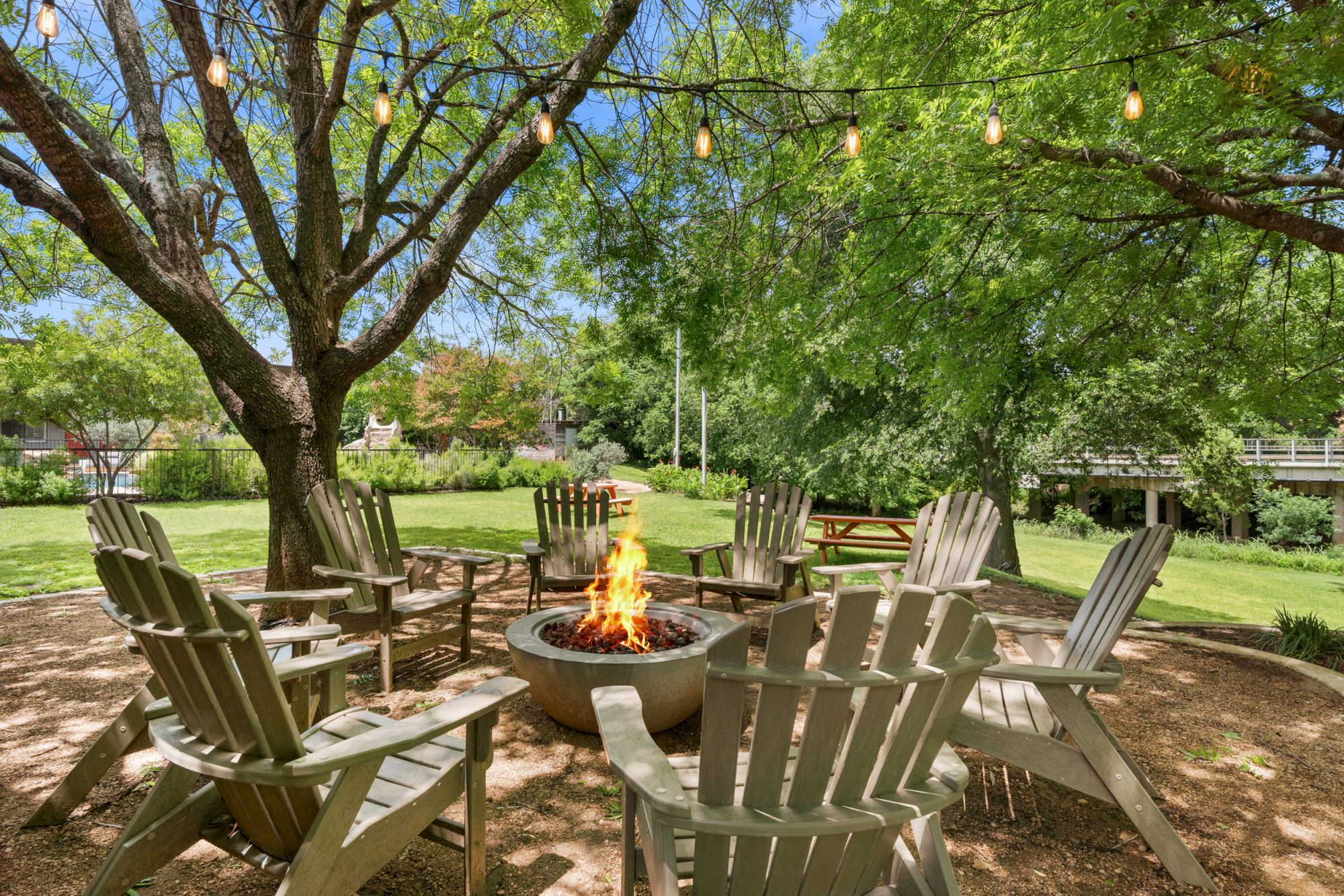 A cozy outdoor fire pit surrounded by wooden chairs under trees with string lights, perfect for a relaxing gathering by the flames.