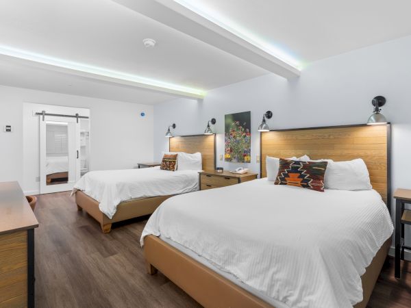A modern hotel room with two queen beds, wooden headboards, white linens, nightstands, a desk, and bright ceiling lighting, spacious and clean.
