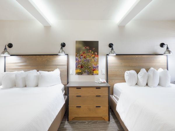 The image shows a modern hotel room with two beds, wooden headboards, bedside lamps, and a colorful painting above a central nightstand.