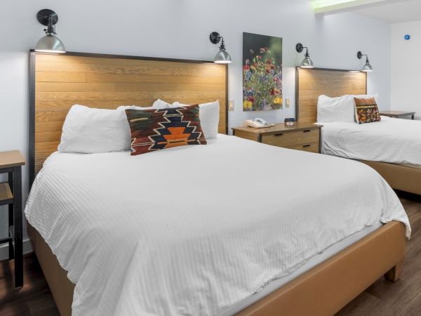 Two neatly made hotel beds with white linens, wooden headboards, matching nightstands, and angled wall lamps in a bright, modern room.
