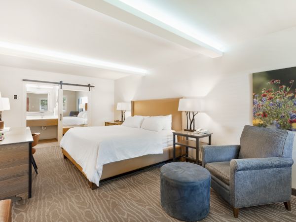 The image shows a hotel room with a bed, bedside tables, a dresser, an armchair, and a small table, decorated in neutral tones.