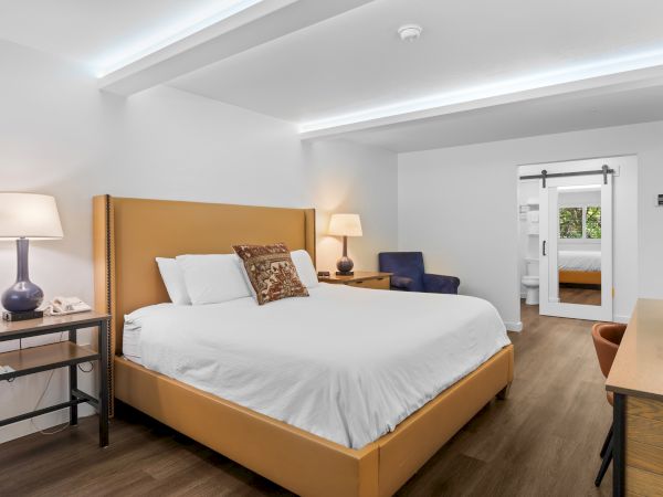 A bright hotel room with a large bed, nightstands, lamps, and a doorway to an adjacent room, modern and tidy.