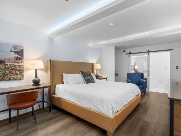 A modern hotel room with a comfy bed, nightstands, lamps, a desk and chair, a blue armchair, and artwork on the wall, bright and inviting.