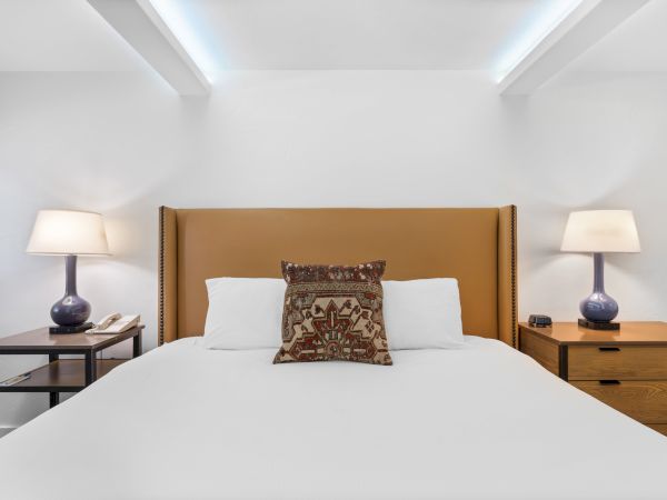 A tidy bedroom with a large bed, tan upholstered headboard, two nightstands with lamps, and a decorative pillow on white bedding.