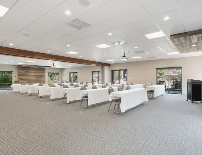 A spacious conference room with neatly arranged tables and chairs, a podium, flags, and large windows letting in natural light.