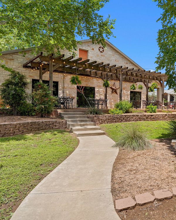 A single-story building with a stone fa&ccedil;ade, a wooden pergola over the entrance, a curved concrete path, landscaped yard, and green trees under a sunny sky.