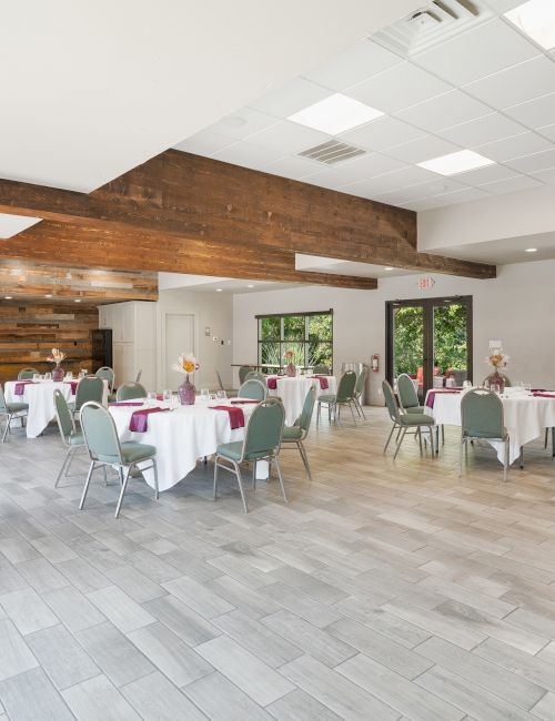 A spacious, brightly lit room with several round tables set for an event, featuring white tablecloths and green chairs, surrounded by windows.