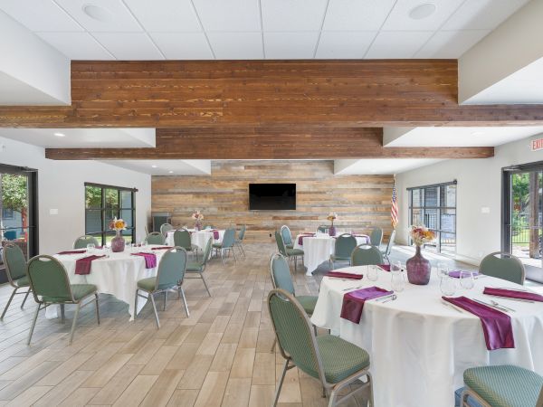 A bright event hall with round tables, white tablecloths, purple napkins, green chairs, and a wood-paneled wall with a mounted TV.