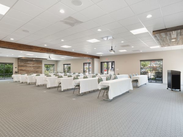 A spacious conference room set up with long, white-draped tables and chairs, American flag, projector, and natural light windows.