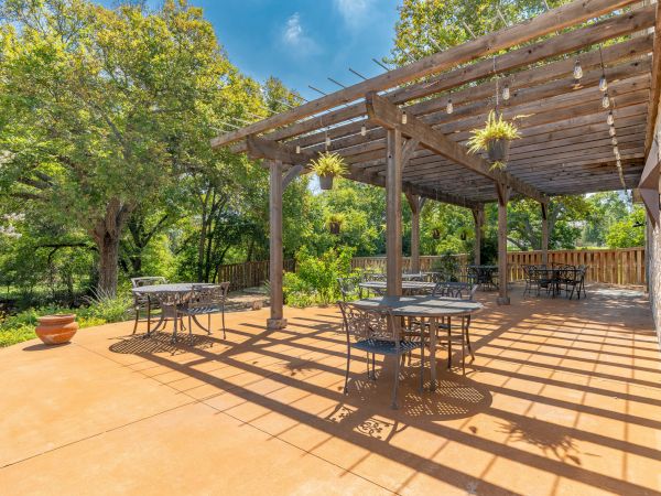 A sunny courtyard porch with a wooden pergola, hanging plants, several metal tables and chairs, and lush green trees in the background.