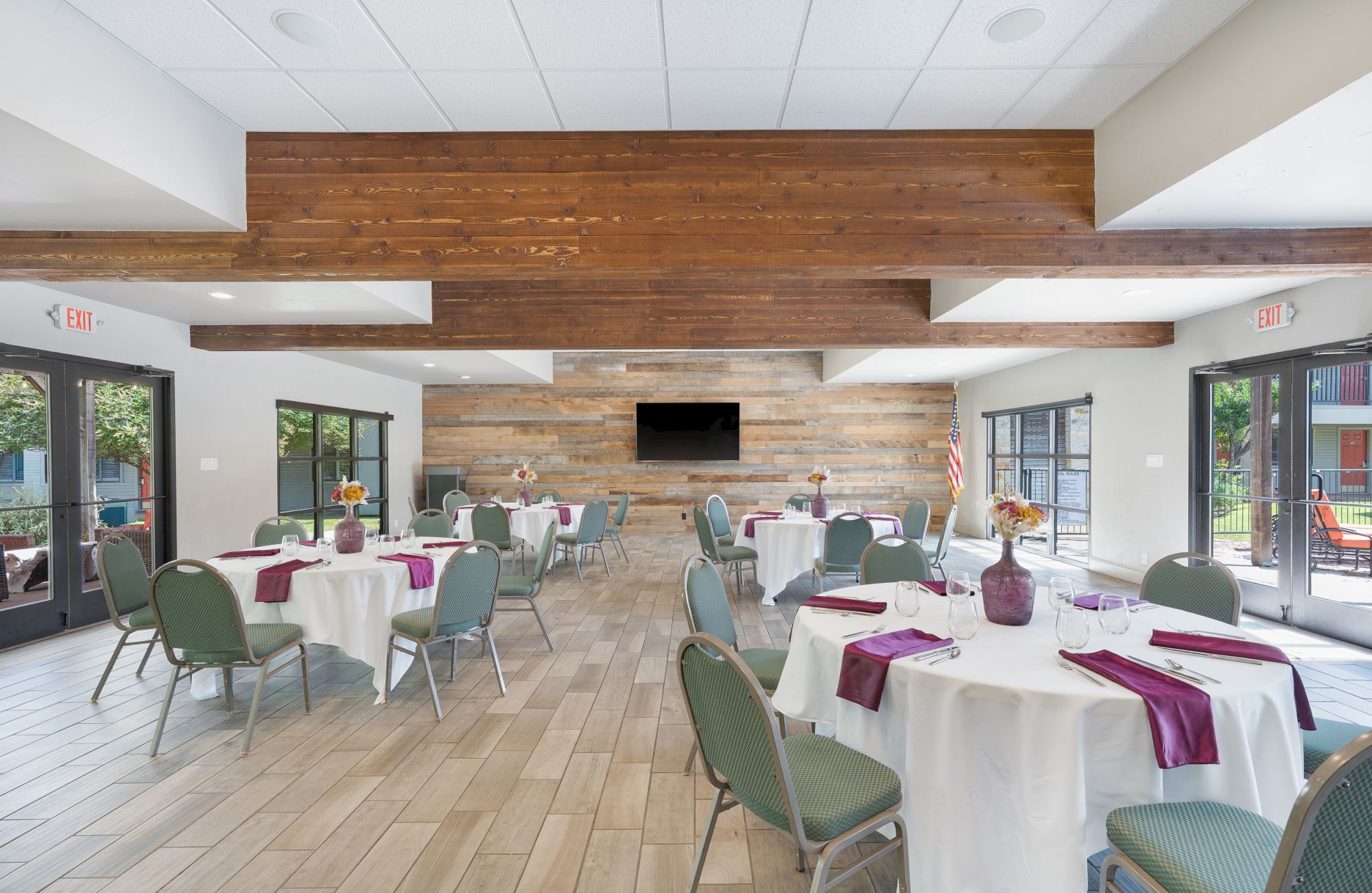 The image shows a modern dining or event room with round tables, green chairs, and a wall-mounted TV, featuring natural light from large windows.