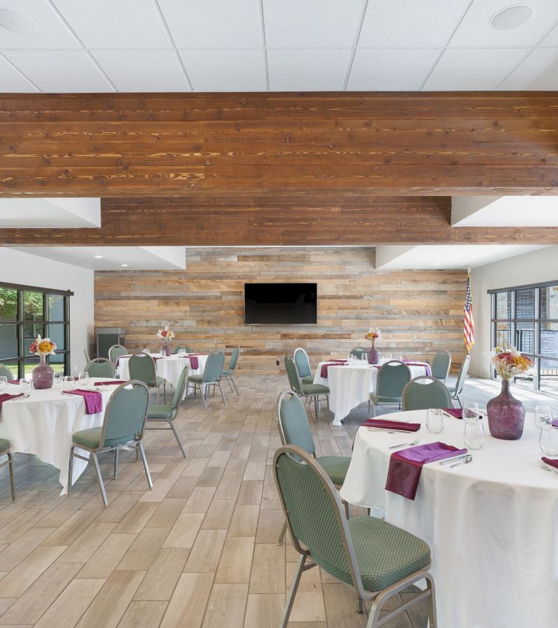 a meeting room with round tables set for dining, a TV on the wall, and windows on either side