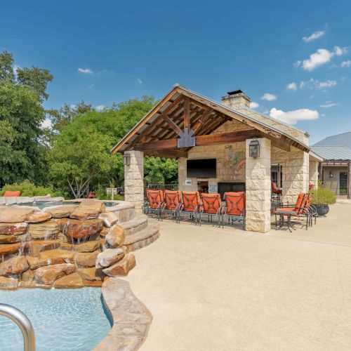 A stone poolside pavilion with a small bar/ grilling area, outdoor seating, a cascading rock waterfall into the pool, and sunny blue skies.