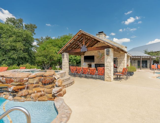 A stone poolside pavilion with a small bar/ grilling area, outdoor seating, a cascading rock waterfall into the pool, and sunny blue skies.