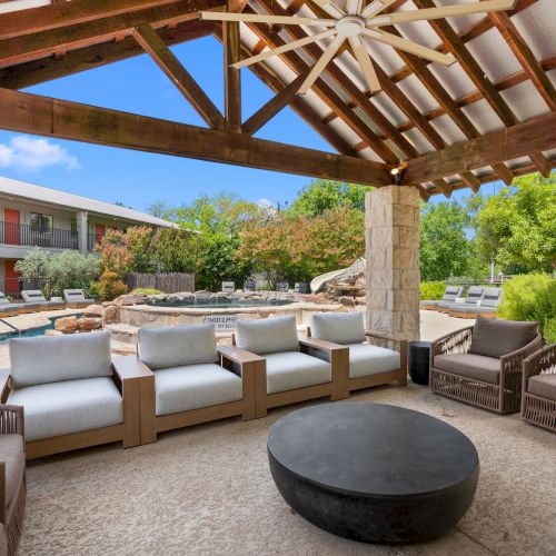 Outdoor covered patio with wicker seating, cushy sofas, and a large round fire pit; stone columns, wooden beams, pool area beyond.