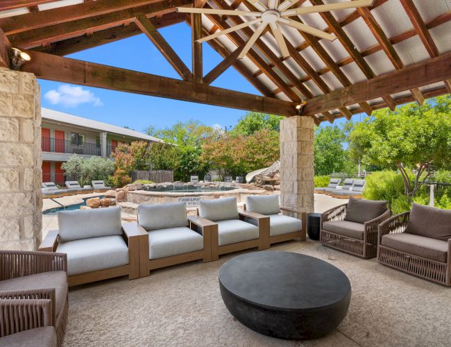 Outdoor covered patio with wicker seating, cushy sofas, and a large round fire pit; stone columns, wooden beams, pool area beyond.