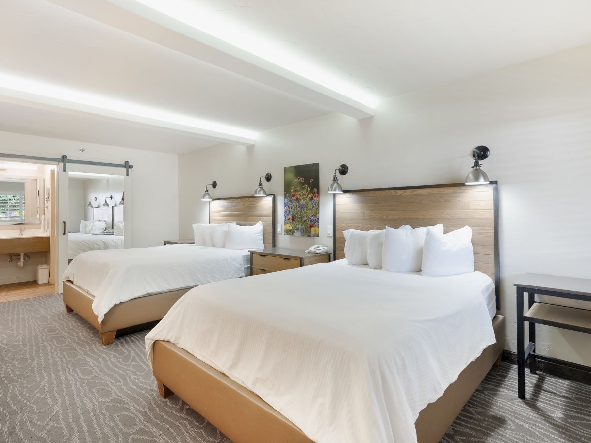 The image shows a hotel room with two double beds, modern decor, and a bathroom in the background.
