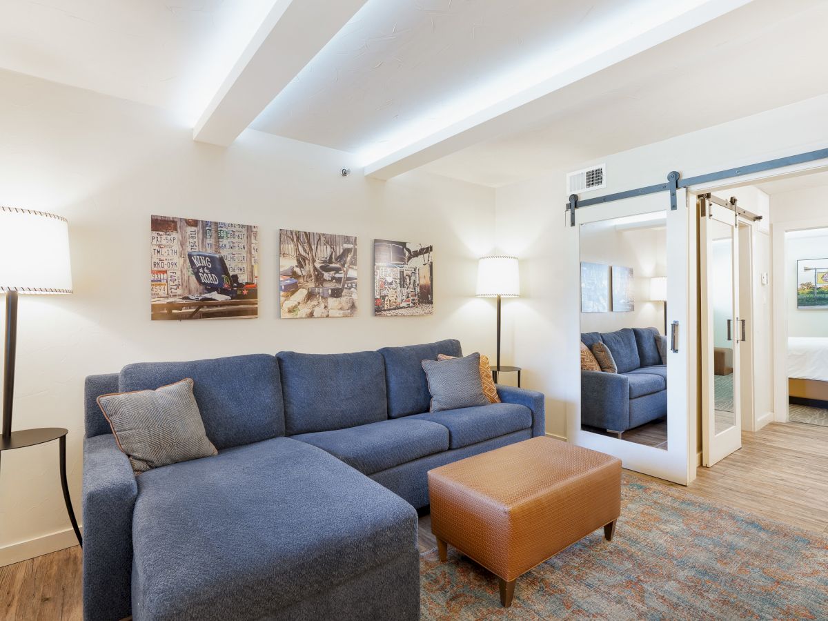 The image shows a cozy living room with a blue sofa, a brown ottoman, two standing lamps, and wall art, leading to a bedroom with a sliding door.