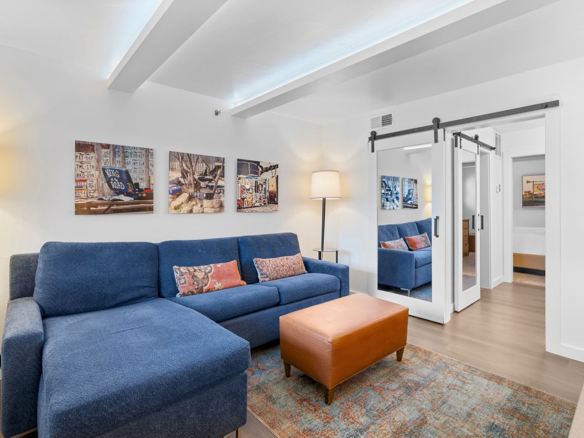 A cozy living room with a blue sectional, orange ottoman, wall art, and sliding barn doors reflecting in mirrored closet doors.