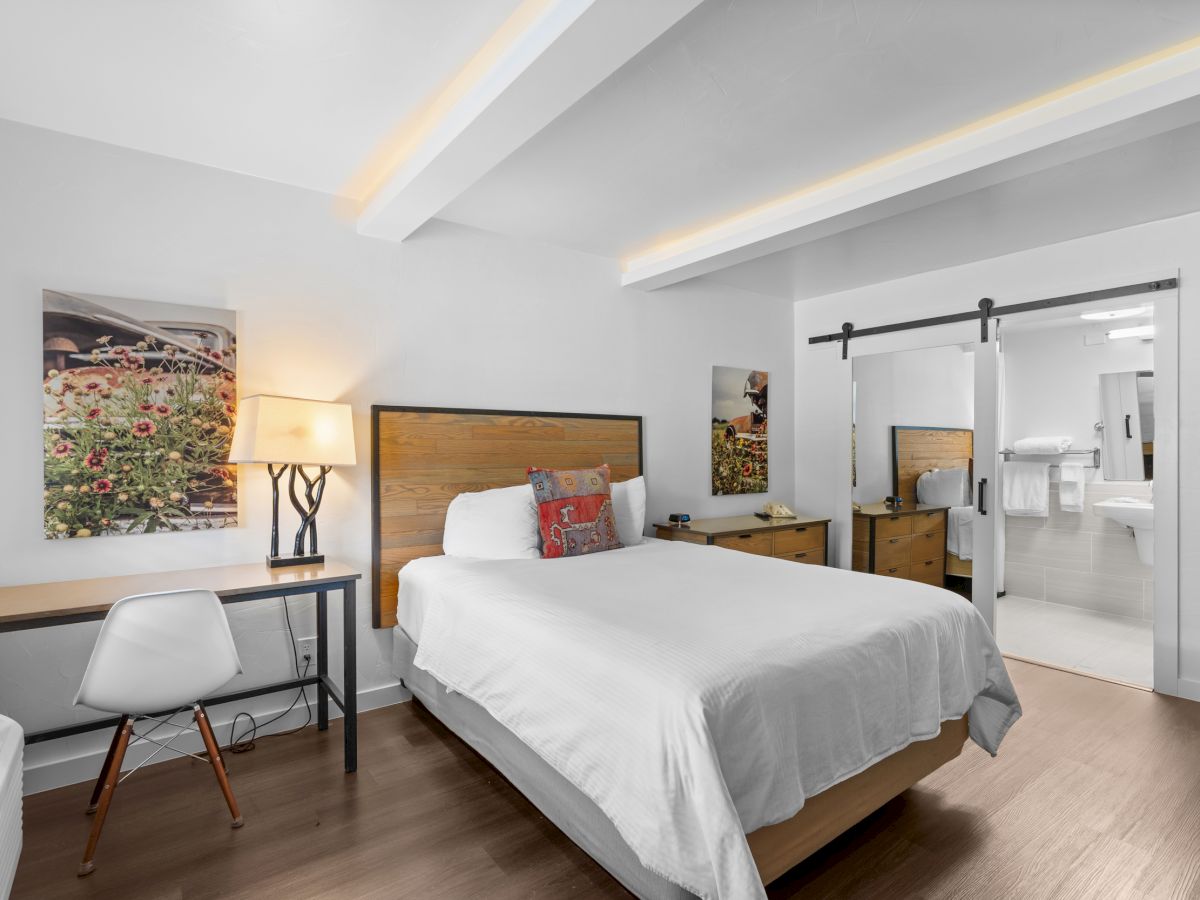 A modern hotel room with a white bed, wooden headboard, desk and chair, lamp, art on the walls, and a sliding barn-door closet leading to a bright adjoining space.