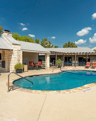 A backyard features a pool surrounded by seating areas and a covered patio under a clear blue sky with scattered clouds.