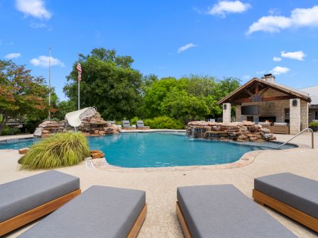 A sunny resort pool area with a curvy blue pool, lounge chairs in the foreground, rock features, a small building, trees, and a clear sky.