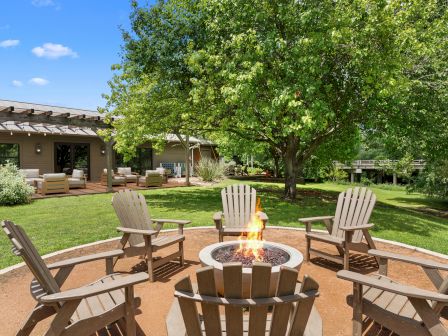 A sunny backyard with a circular fire pit surrounded by wooden chairs, a lush lawn, a big tree, and a house patio in the background.