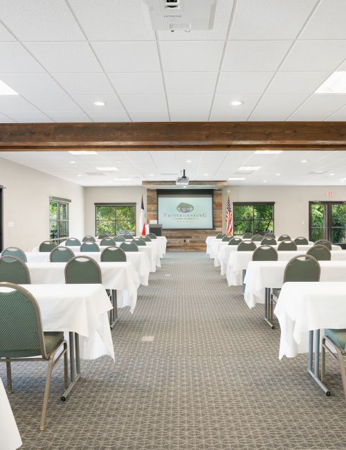 The image shows a conference room setup with rows of tables and chairs, a projection screen, and natural light from large windows.