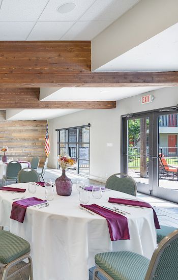 A dining area with round tables set for a meal, featuring white tablecloths, purple napkins, and vases with flowers.
