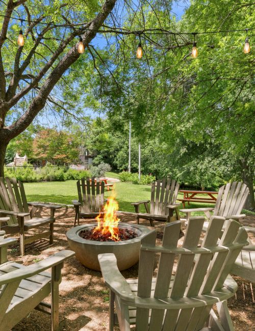 A cozy outdoor seating circle around a fire pit under trees with string lights and wooden chairs, perfect for conversations and warmth at night.