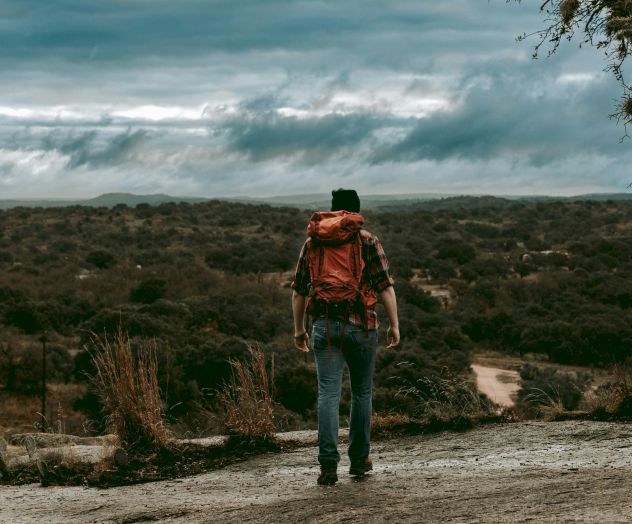 A person stands on a dirt road in a hilly landscape with a cloudy sky, wearing a red jacket and backpack, looking at the camera.