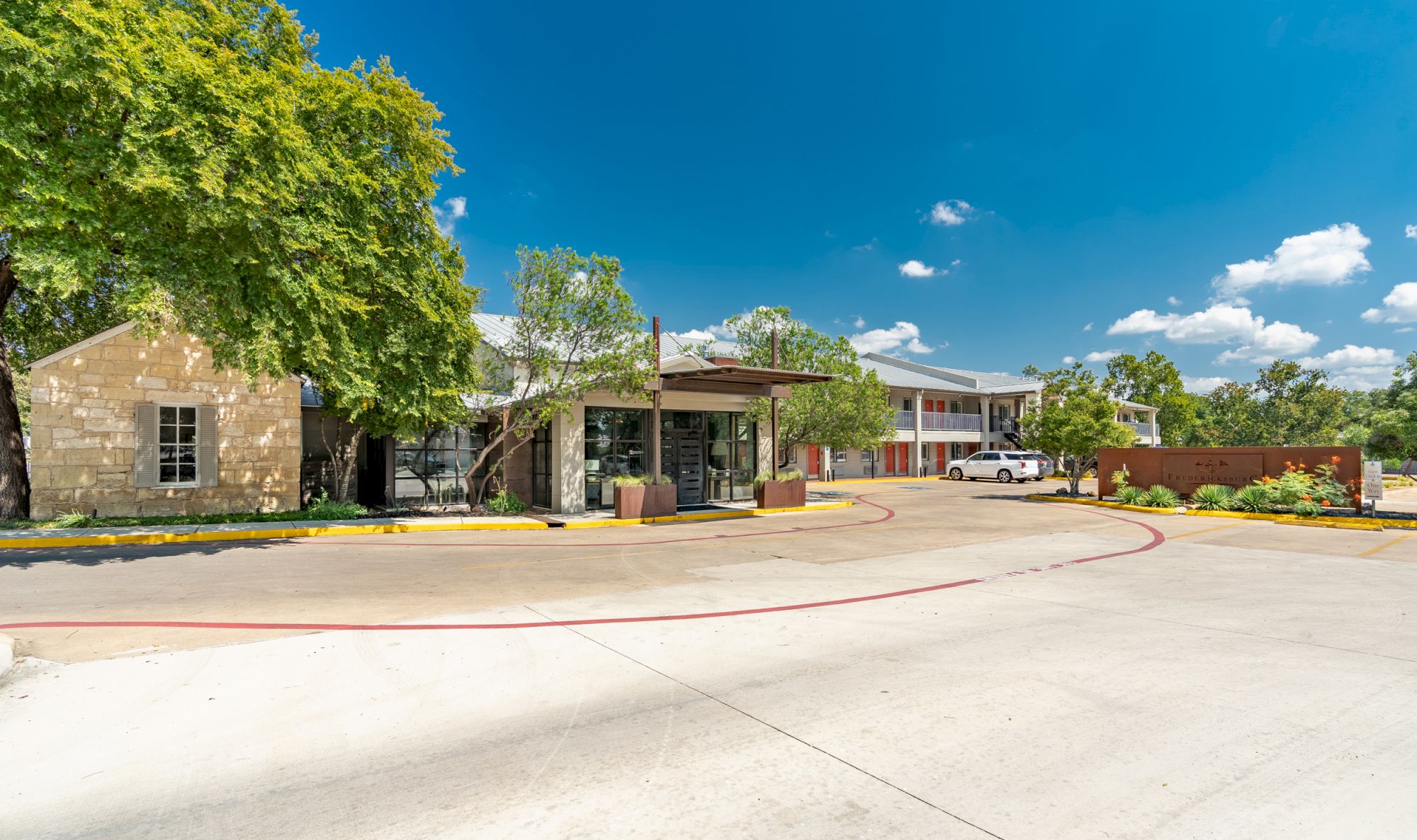 The image shows a sunny day at a rustic building with trees, a pathway, and a parking area, set against a blue sky with clouds.