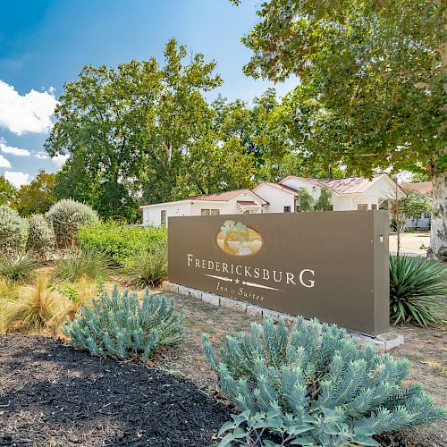 The image shows a landscaped area with a sign for Fredericksburg, featuring greenery and clear blue sky.