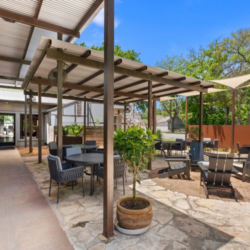 Outdoor patio with a covered pergola, lounge chairs, small tables, potted plants, and a bright blue sky.