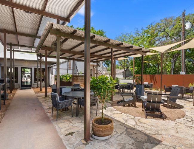 Outdoor patio with a covered pergola, lounge chairs, small tables, potted plants, and a bright blue sky.