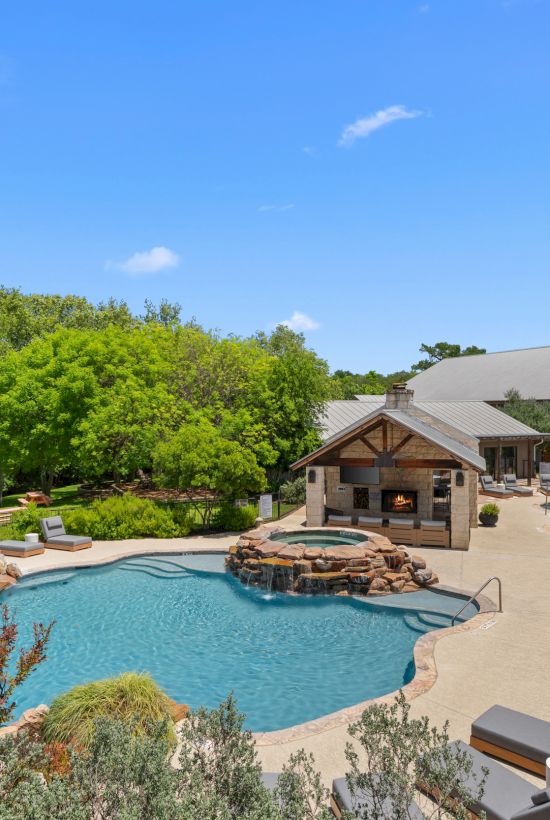 Outdoor resort pool with waterfall, sun loungers, firepit, stone deck, and a large barn-style building under a blue sky.