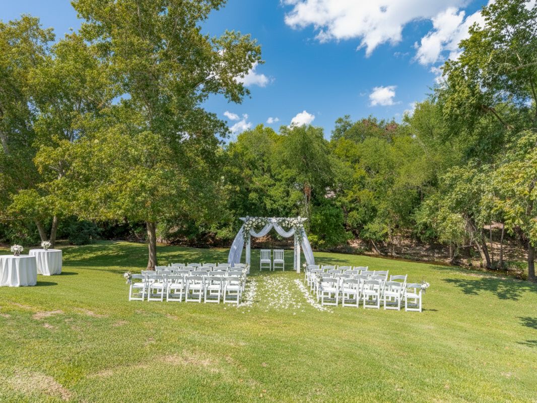A beautifully decorated outdoor event space with a white archway, chairs, and flowers on a grassy area surrounded by trees.