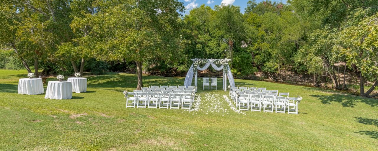 An outdoor wedding ceremony setup with white chairs, tables, and an archway in a grassy area with trees.