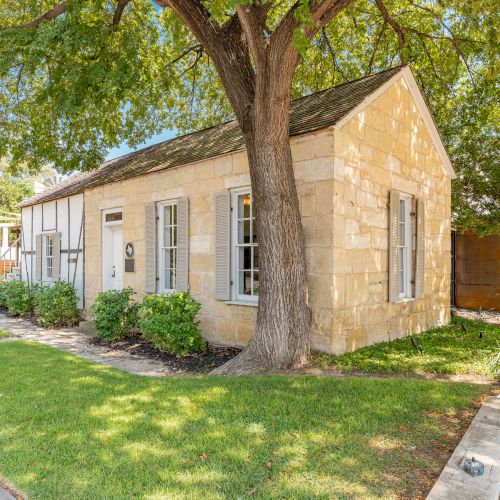 A small stone house with a gabled roof is surrounded by grass and a large tree in front, alongside a sidewalk.