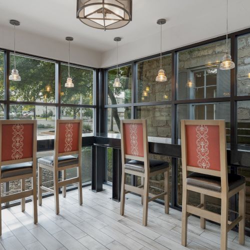 A modern dining area with high chairs and tables, large windows, pendant lights, and a view of outdoor greenery and stone walls.