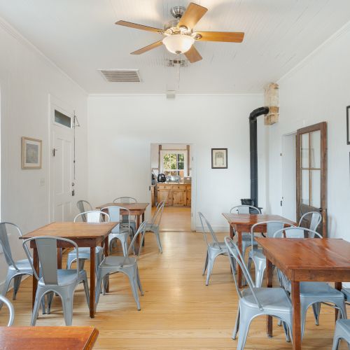 The image shows a bright room with wooden tables, metal chairs, a ceiling fan, artwork on the walls, and large windows.