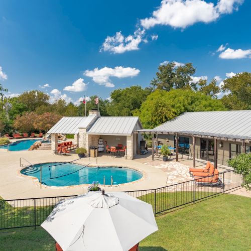 A backyard features a swimming pool, a shaded seating area, and a white umbrella on a sunny day with a blue sky and scattered clouds.