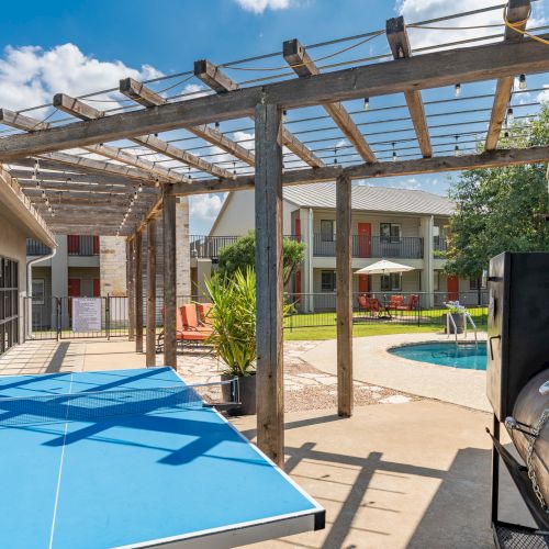 The image shows a backyard with a pergola, a table tennis table, a barbecue grill, and a swimming pool in a sunny, residential setting.