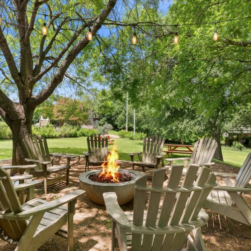 A sunny backyard patio with a crackling fire pit, Adirondack chairs, string lights, and lush green trees surrounding a grassy lawn.